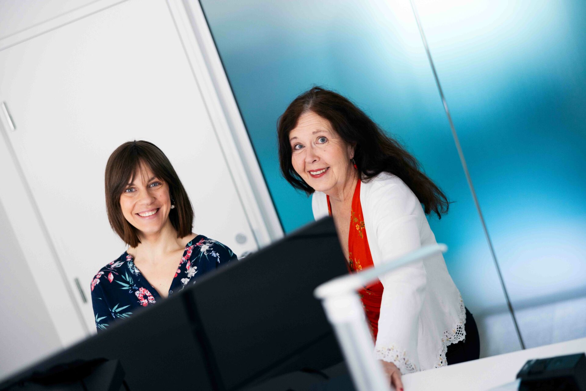 Kaksi hymyilevää naista toimistolla koneen äärellä. Katsovat kameraan.