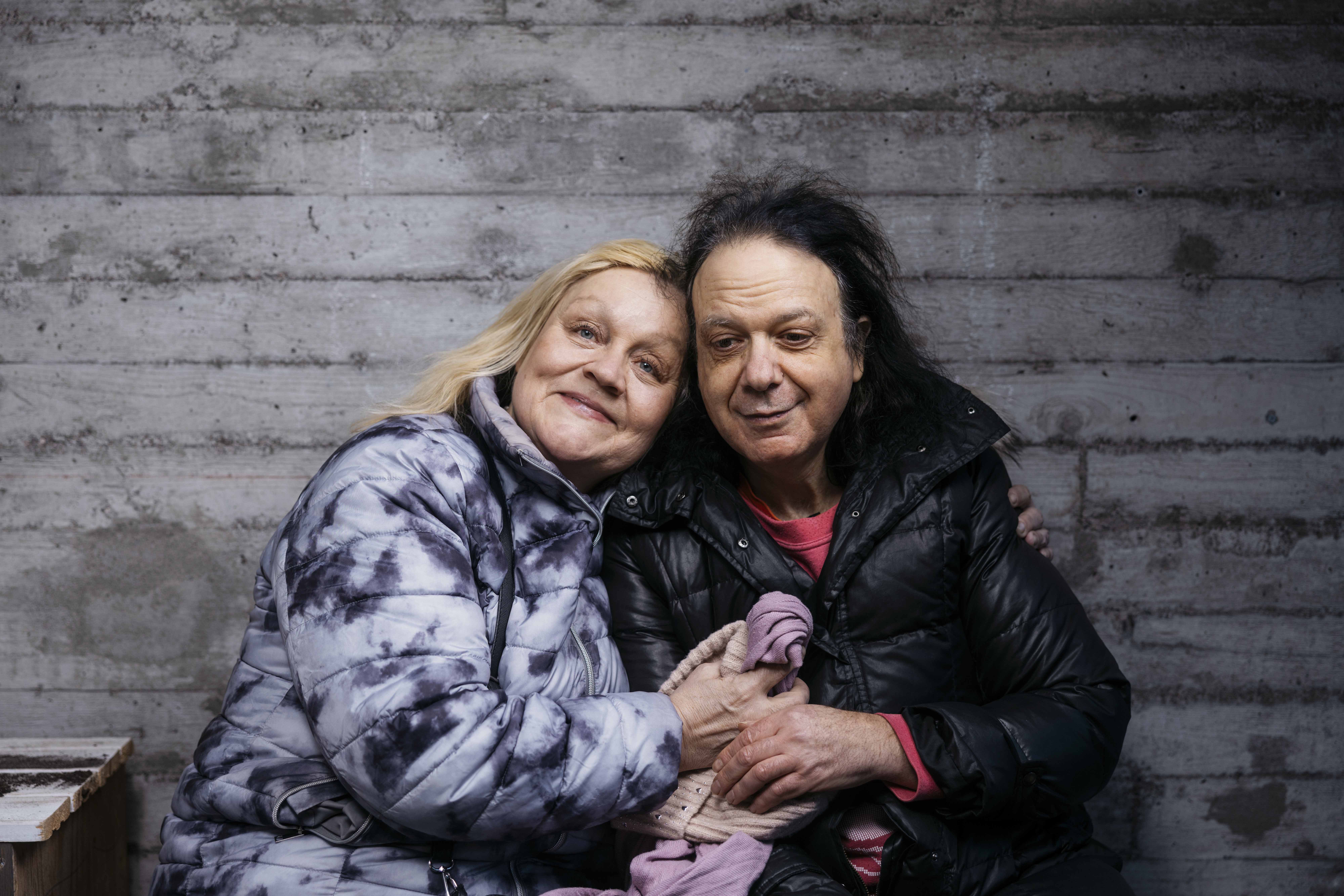 Tummahiuksinen mies ja vaalehiuksinen nainen toppatakeissa, nojaavat toisiinsa, noin 60-vuotiaita. Toiveikkaat ilmeet.