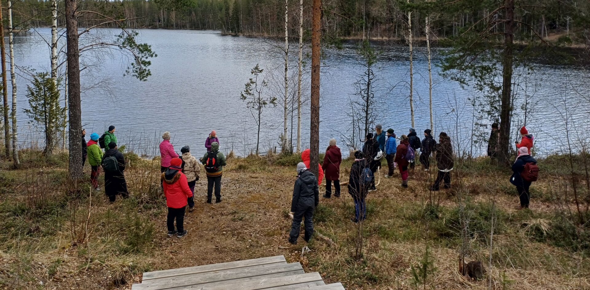 Ihmisiä järven rannalla retkivaatetuksessa. Alkukevään päivä.