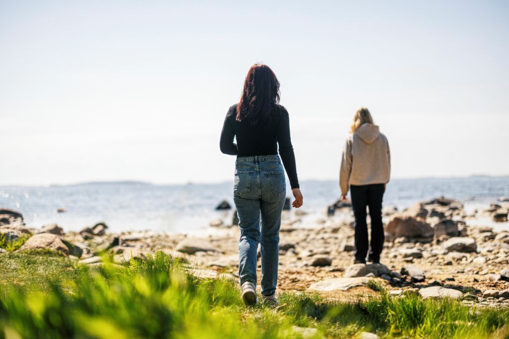 Kaksi nuorta meren rannalla, selin kameraan. Tummahiuksinen nuori tyttö kävelee vaaleahiuksisen tytön luo.