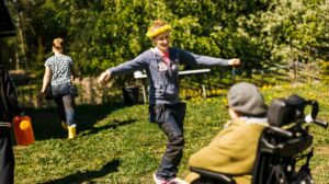 Noin 40-vuotiaalla naisella on keltainen kukkaseppele päässään ja kädet sivulla. Hän katsoo pyörätuolissa olevaa vanhempaa naista, joka on selin. Kesäinen maisema.