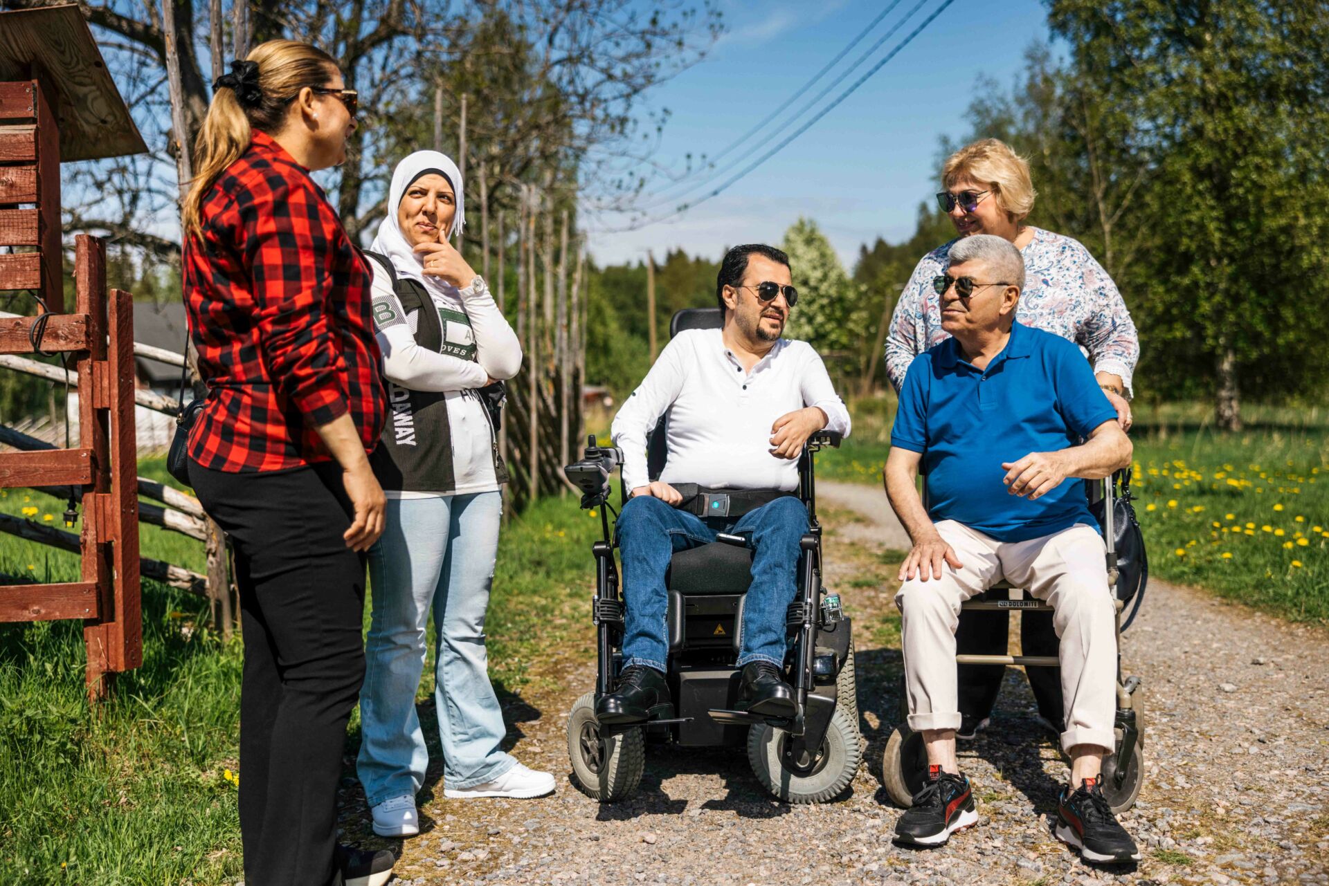 Viisi ihmistä kesäisellä hiekkatiellä maalaismaisemassa. Kolme naista seisoo joista yhdellä on valkoinen hijab ja kaksi miestä istuu pyörätuoleissa.
