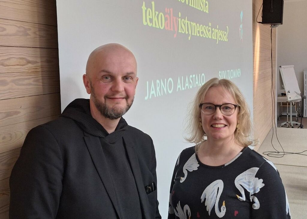 Hymyilevä, kalju, parrakas mies, jolla on musta pikkutakki. Hymylevä, vaalea, silmälasipäinen nainen, jolla on polkkatukka ja musta-valkea pusero.