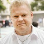 Sänkitukkainen, vakava mies, jolla on kauluspaita ja kaulassa ristiriipus. Taustalla maisemaa.