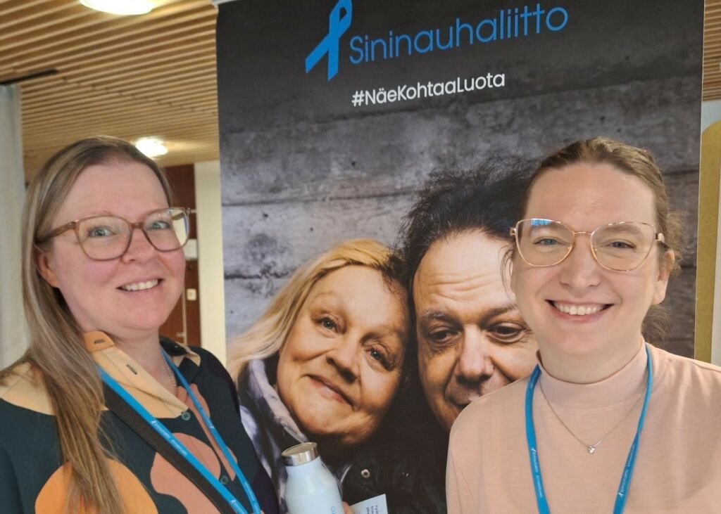 Pitkätukkainen, silmälasipäinen nainen ja lyhyttukkainen silmälasipäinen nainen hymyilevät leveästi Sininauhaliiton roll-upin edessä. Roll-upissa on kaksi henkilöä, jotka nojaavat toisiinsa, ja teksti Sininauhaliitto Näe Kohtaa Luota.